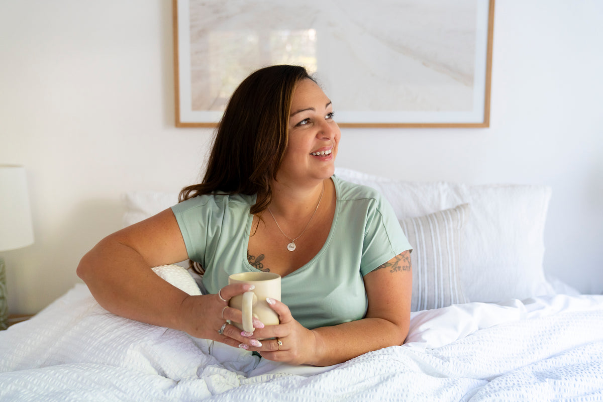 Happy woman looking comfortable and cool in a white fresh linen bed wearing soft silky pyjamas for night sweats and holding a coffee mug
